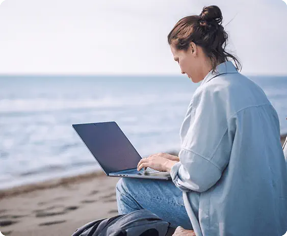 Mujer sentada a la orilla del mar escribiendo en su portátil