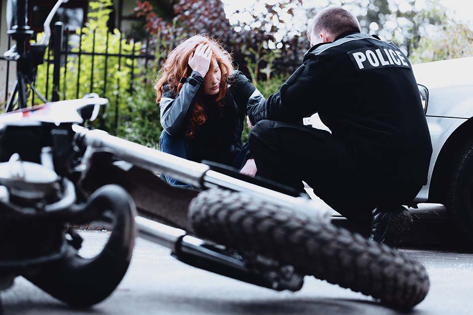 Motorista sentada en el suelo con las manos en la cabeza tras haber tenido un accidente de tráfico. Policía en cuchillas a su lado realizando el atestado policial